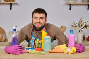 giovane bello barbuto uomo nel il cucina, Spettacoli tutti il suo pulizia personale - detersivi, spazzole, spray. lui pensare lui è pronto per vero pulizia foto