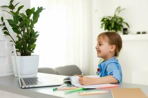 inteligente poco ragazza fa compiti a casa nel sua vivente camera. lei è seduta a sua scrivania scrive con un' penna nel sua libri di testo e usi il computer portatile. foto