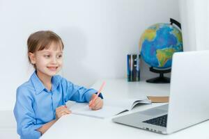 carino poco ragazza è seduta a tavolo con sua il computer portatile e studiando in linea foto