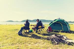 gruppo di viaggiatori che si accampano e fanno picnic e suonano musica insieme. sfondo di montagna e lago. persone e stile di vita. attività all'aperto e tema del tempo libero. zaino in spalla ed escursionista. angolo di visione posteriore foto