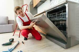 tecnico o lavoratore nel uniforme installa lavastoviglie in il cucina arredamento. riparatore indossare lavoratore completo da uomo riparazione Manutenzione di lavastoviglie. maestro nel protettivo guanti fix lavastoviglie. foto