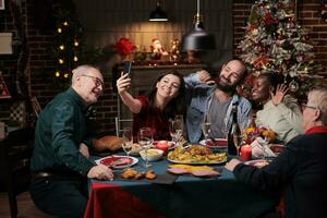 famiglia assunzione fotografie a Natale cena con delizioso cibo e bicchieri di vino, fabbricazione ricordi durante dicembre vacanza evento. diverso persone avendo divertimento con immagini su smartphone a casa.