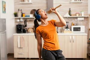 cantando su di legno cucchiaio indossare cuffie nel il cucina. energico, positivo, felice, divertente e carino casalinga danza solo nel il Casa. divertimento e tempo libero solo a casa. foto