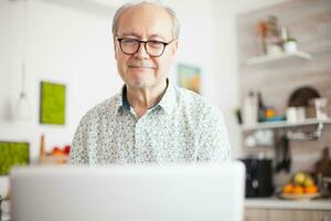 pensionato uomo sorridente mentre Guardando un' film su il il computer portatile. quotidiano vita di anziano uomo nel cucina durante prima colazione utilizzando il computer portatile Tenere un' tazza di caffè. anziano pensionato persona Lavorando a partire dal casa, telelavoro utilizzando a distanza Internet lavoro in linea comunicazione. foto