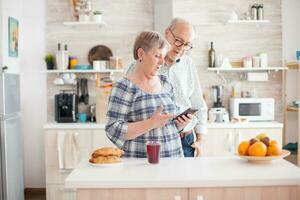 caucasico anziano moglie e marito nel cucina durante prima colazione utilizzando smartphone tecnologia e Internet connessione. tempo libero pensionato donna e uomo sorridente nel domicilio, rilassante. foto