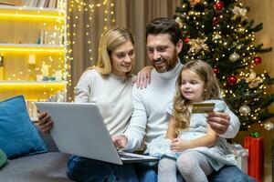 contento Natale famiglia fare in linea shopping seduta a casa su divano, coppia marito moglie e figlia con il computer portatile e banca credito carta avendo divertimento la scelta Natale i regali vicino Natale albero. foto