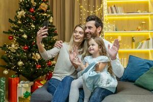 contento famiglia festeggiare nuovo anno e Natale, marito moglie e poco figlia seduta su divano nel vivente camera e parlando con parenti su video chiamata, utilizzando Telefono per a distanza comunicazione. foto