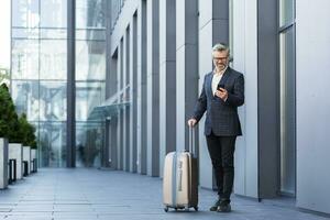 un' uomo nel un' attività commerciale completo da uomo con un' grande viaggio valigia e un' Telefono è digitando un' Messaggio e prenotazione un' Taxi, un' capo su un' attività commerciale viaggio, un' maturo uomo d'affari al di fuori un' grande Hotel. foto