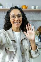 giovane bellissimo ispanico donna nel bicchieri e beige camicia scrive un' blog, conduce un in linea flusso. parlando per il telecamera a casa nel il cucina. lei sorrisi felicemente e onde sua mano. foto