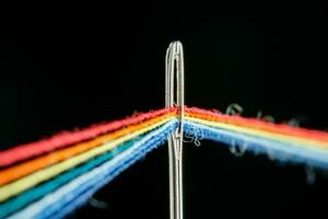 multicolore discussioni per cucire nel il modulo di un' arcobaleno passaggio attraverso un antico ago su un' nero sfondo foto