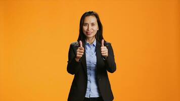 filippina dipendente fa pollici su gesto su telecamera, esprimendo positività con piace simbolo al di sopra di arancia sfondo. giovane donna nel formale abbigliamento presentazione approvazione cartello nel studio. foto