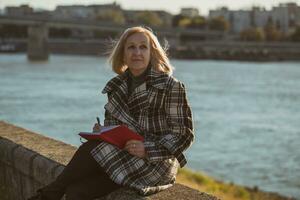 anziano donna gode scrittura qualcosa mentre seduta di il fiume.tonico Immagine. foto