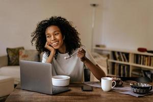 giovane donna nera con gli auricolari che usa il laptop e fa colazione? foto