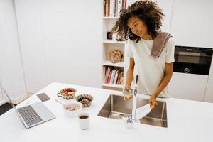giovane donna nera che lava i piatti mentre usa il laptop in cucina foto