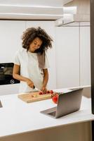 giovane donna nera che prepara insalata mentre usa il computer portatile in cucina foto