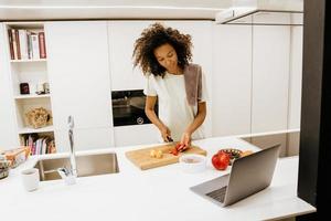 giovane donna nera che prepara insalata mentre usa il computer portatile in cucina foto
