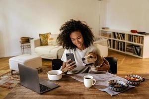 donna di colore che usa il cellulare e abbraccia il suo cane mentre fa colazione foto