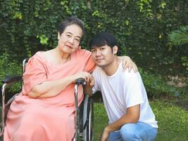 asiatico anziano donna seduta su sedia a rotelle con sua figlio nel il giardino. foto