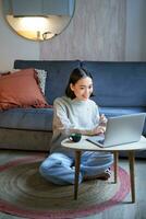 verticale tiro di asiatico ragazza si siede su pavimento a casa, Lavorando su computer portatile, studiando a accogliente posto, utilizzando computer per libero professionista foto