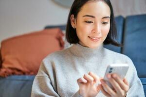 ritratto di sorridente asiatico donna si siede a casa, usi smartphone app, ordini consegna, messaggi qualcuno su mobile Telefono applicazione foto