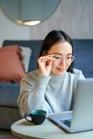 giovane asiatico ragazza, libero professionista professionale seduta nel vivente camera e Lavorando con computer, studiando a casa utilizzando computer foto