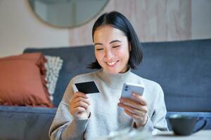 ritratto di sorridente asiatico donna pagare con credito carta su sua smartphone app, organizzare diretto addebito o shopping in linea a partire dal mobile Telefono foto