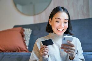 ritratto di sorridente asiatico donna pagare con credito carta su sua smartphone app, organizzare diretto addebito o shopping in linea a partire dal mobile Telefono foto