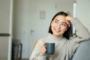 sorridente asiatico donna seduta su divano con sua tazza, potabile caffè a casa e rilassante dopo opera, guardare calma e accogliente foto