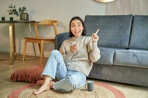 ritratto di elegante coreano donna si siede su pavimento con smartphone, utilizzando mobile Telefono, sorridente lieto, concetto di soggiorno a casa e rilassamento foto