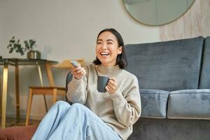 ritratto di bellissimo asiatico ragazza seduta a sua casa e Guardando tv, Tenere a distanza, sorridente e ridendo, sensazione comfort e calore a sua appartamento foto
