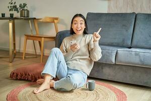 sorridente asiatico ragazza si siede su pavimento nel elegante vivente camera, puntamento dito a annuncio, mostrando promo striscione, Tenere mobile Telefono nel mano foto