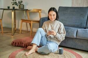 Immagine di elegante giovane donna nel moderno Casa, utilizzando mobile Telefono, seduta su pavimento e Tenere smartphone, potabile a partire dal tazza foto