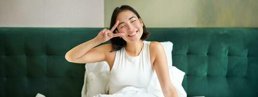 positivo asiatico donna dire bugie nel letto, mostrando pace cartello, gode contento mattina, veglia su ottimista, soggiorno nel sua Camera da letto foto
