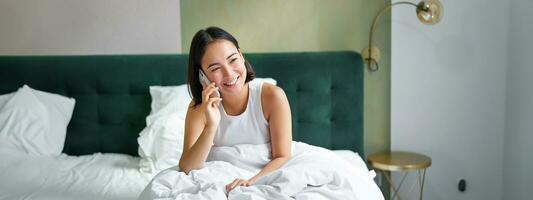 bellissimo giovane coreano donna chiamando, avendo un' Telefono chiamata nel letto, parlando con amico mentre soggiorno nel Camera da letto foto