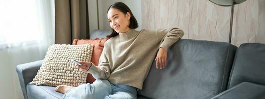 bellissimo asiatico ragazza nel accogliente casa, Guardando televisione, Tenere a distanza e commutazione canali, seduta su divano foto