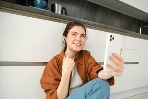 ritratto di contento sorridente donna video chat, si siede su cucina pavimento a casa, parlando per amico su sociale media App utilizzando smartphone foto