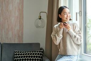 ritratto di bellissimo asiatico donna seduta a casa con tazza di caffè, godendo sua caffè espresso mentre guardare al di fuori finestra a passante, sorridente felicemente foto