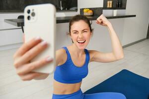 giovane energico ragazza fa gli sport a casa, Spettacoli sua muscoli su braccio e prende autoscatto su smartphone, in posa su fitness gomma da cancellare stuoia nel vivente camera foto