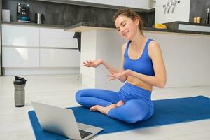 Immagine di giovane sorridente donna, fitness istruttore seduta su yoga stuoia, parlando per cliente attraverso in linea video Chiacchierare su computer portatile, insegnamento aerobica, allenarsi formazione foto