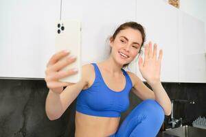 ritratto di sportiva, fitness blogger, registrazione video, agitando mano a smartphone schermo e sorridente, in linea chattare, seduta nel cucina, indossare abbigliamento sportivo foto