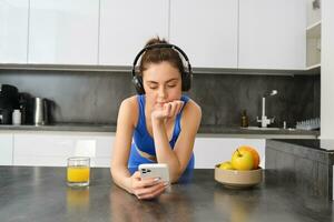 ritratto di fitness ragazza, in piedi nel cucina, ascoltando musica e guardare a smartphone, potabile arancia succo a partire dal bicchiere, indossare abbigliamento sportivo foto