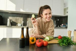 ritratto di contento ragazza mangiare verdure mentre fabbricazione pasto, cucinando salutare vegetariano cibo nel cucina, Tenere zucchine, in posa nel accappatoio foto