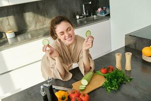 ritratto di bellissimo brunetta donna nel il cucina, indossare accappatoio, chopping verdure su asse, cucinando salutare vegetariano cibo, preparazione insalata, fabbricazione un' pasto foto