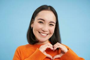 vicino su ritratto di bello asiatico donna, Spettacoli cuore cartello e sorrisi con cura e tenerezza, sta al di sopra di blu sfondo foto