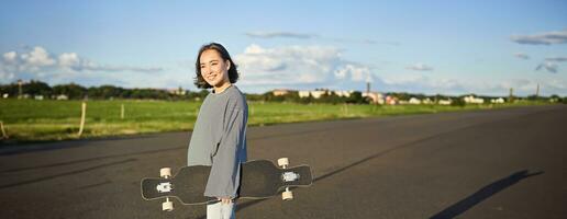 verticale tiro di spensierato asiatico ragazza con longboard. giovane donna pattinatore Tenere incrociatore su sua le spalle e a piedi su strada, andare con lo skateboard foto