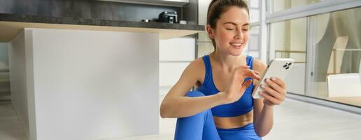 Immagine di giovane in forma donna indossare reggiseno sportivo e blu ghette, utilizzando smartphone app, fa allenarsi a partire dal casa, si siede su pavimento e segue in linea formazione video foto