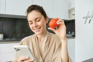 Immagine di sorridente bellissimo donna, si siede nel cucina, detiene smartphone e pomodoro, sembra felice, ordini drogheria consegna per sua casa, utilizzando mobile Telefono app, acquisto in linea verdure per sua ricetta foto