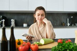 donna Guardando ricetta su smartphone, seduta nel il cucina con verdure e chopping asse, preparazione cena, salutare insalata, cucinando a casa, indossare accappatoio foto