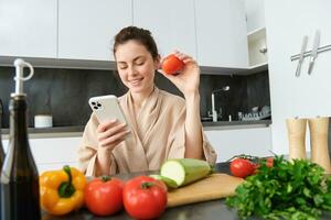 giovane donna ordini drogheria su mobile app. ragazza nel accappatoio si siede nel il cucina con la verdura, guardare per ricetta per cucinare cena, utilizzando smartphone applicazione foto