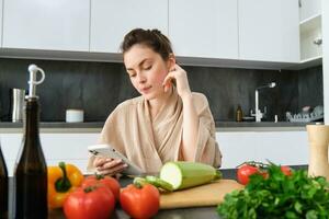 giovane donna ordini drogheria su mobile app. ragazza nel accappatoio si siede nel il cucina con la verdura, guardare per ricetta per cucinare cena, utilizzando smartphone applicazione foto